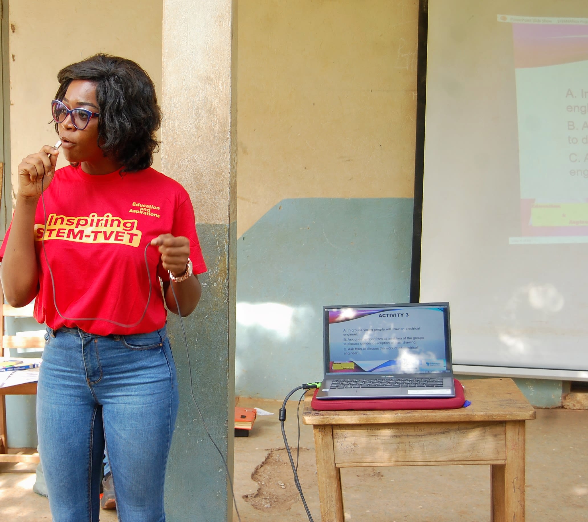 Alice stands holding a microphone. She wears a bright red t-shirt which reads "inspiring STEM-TVET"