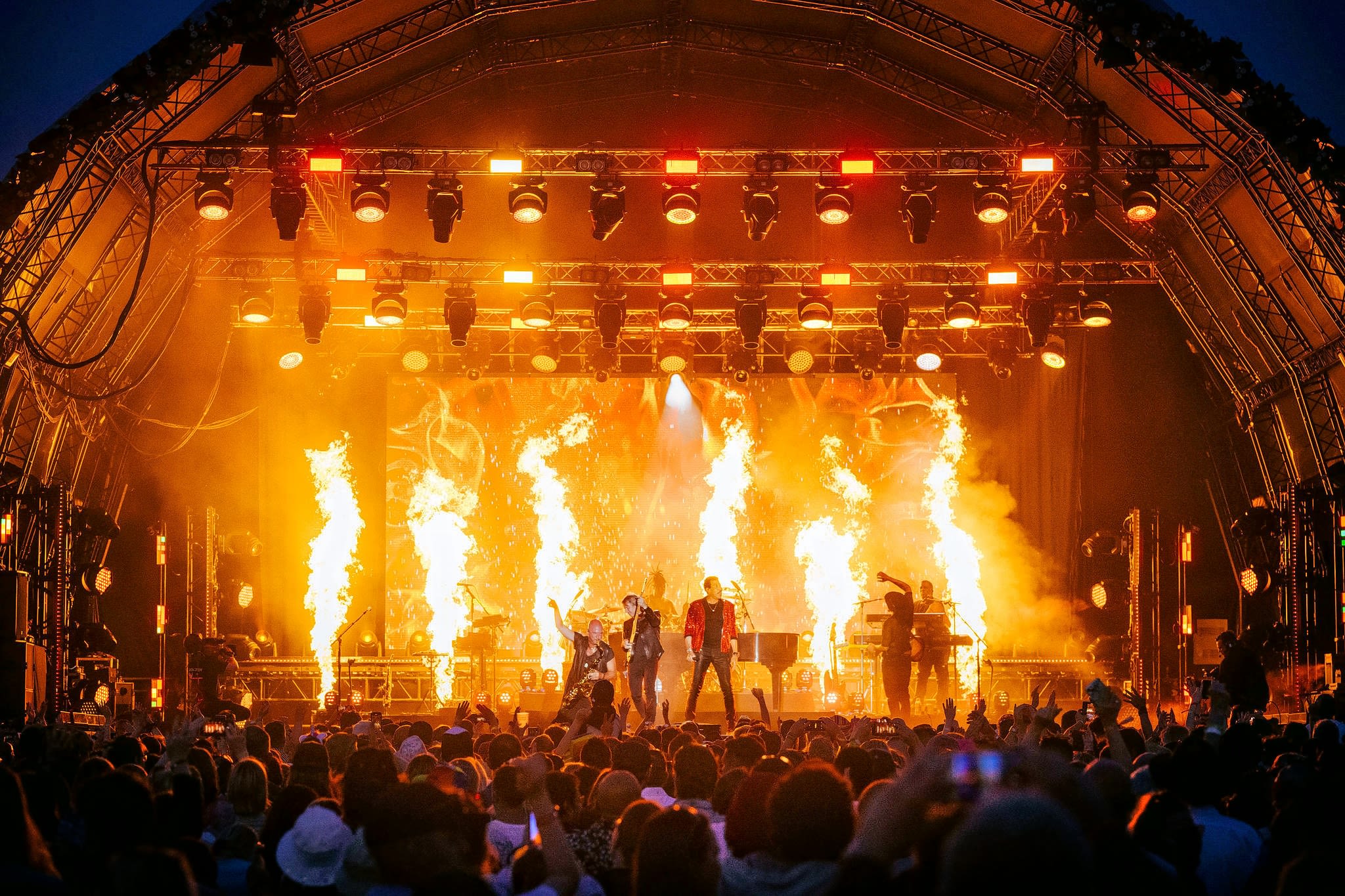 A brightly lit festival stage shows a band silhouetted against a back-drop of orange flames.