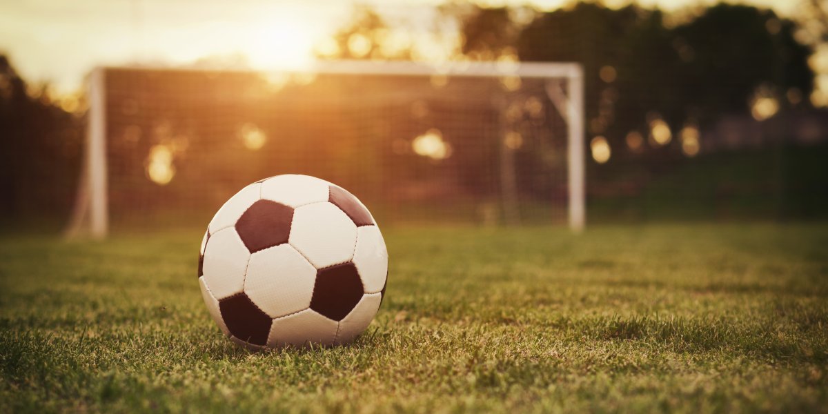 A football sits on a grass pitch while the sun sets in the background of a goal.